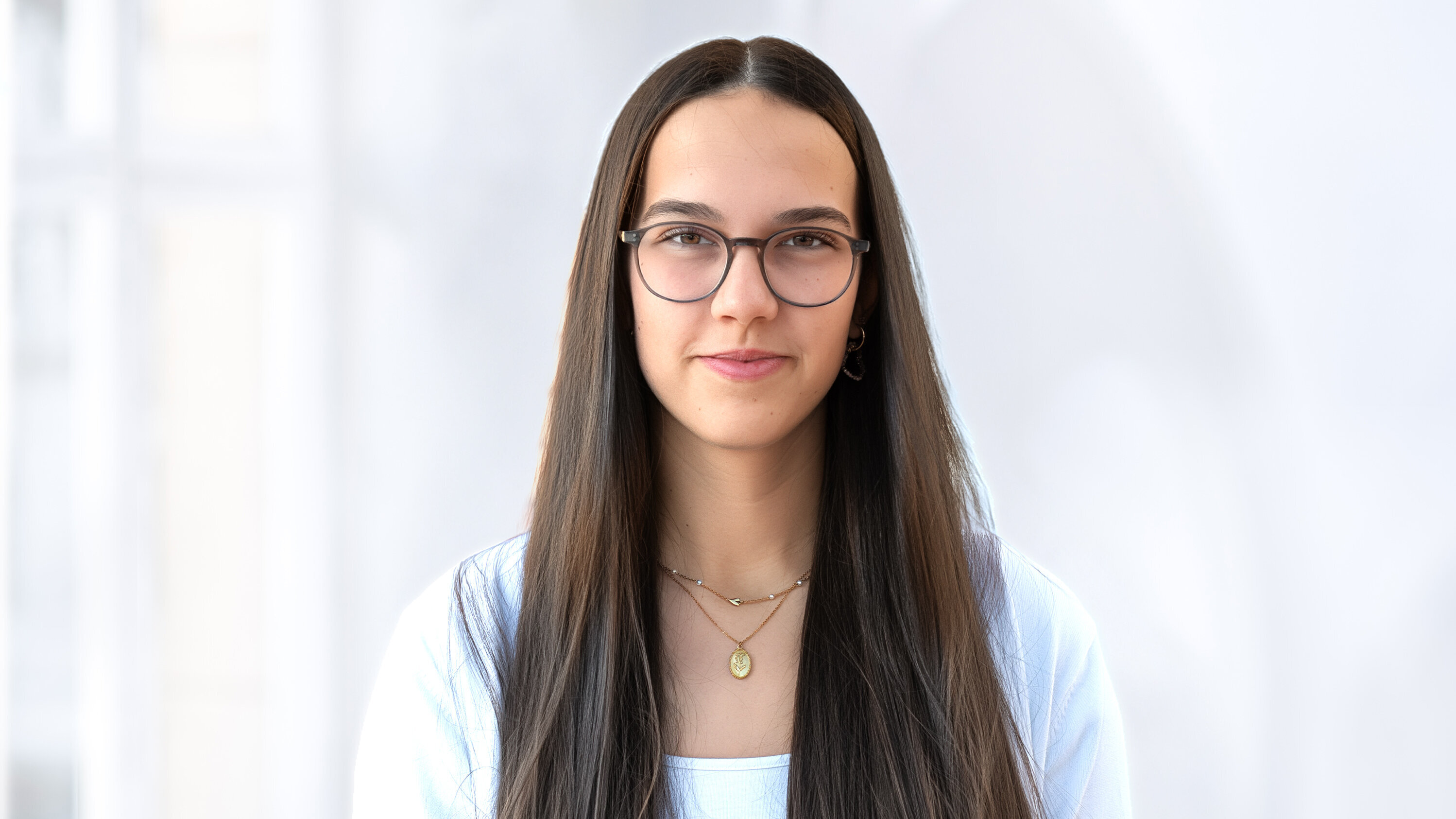 Eine junge Frau mit langen, dunklen Haaren steht vor einem hellen Hintergrund. Sie trägt eine weiße Bluse und eine goldene Halskette. Ihre Brille hat einen zarten, modischen Rahmen. Ihr Ausdruck ist freundlich und selbstbewusst.