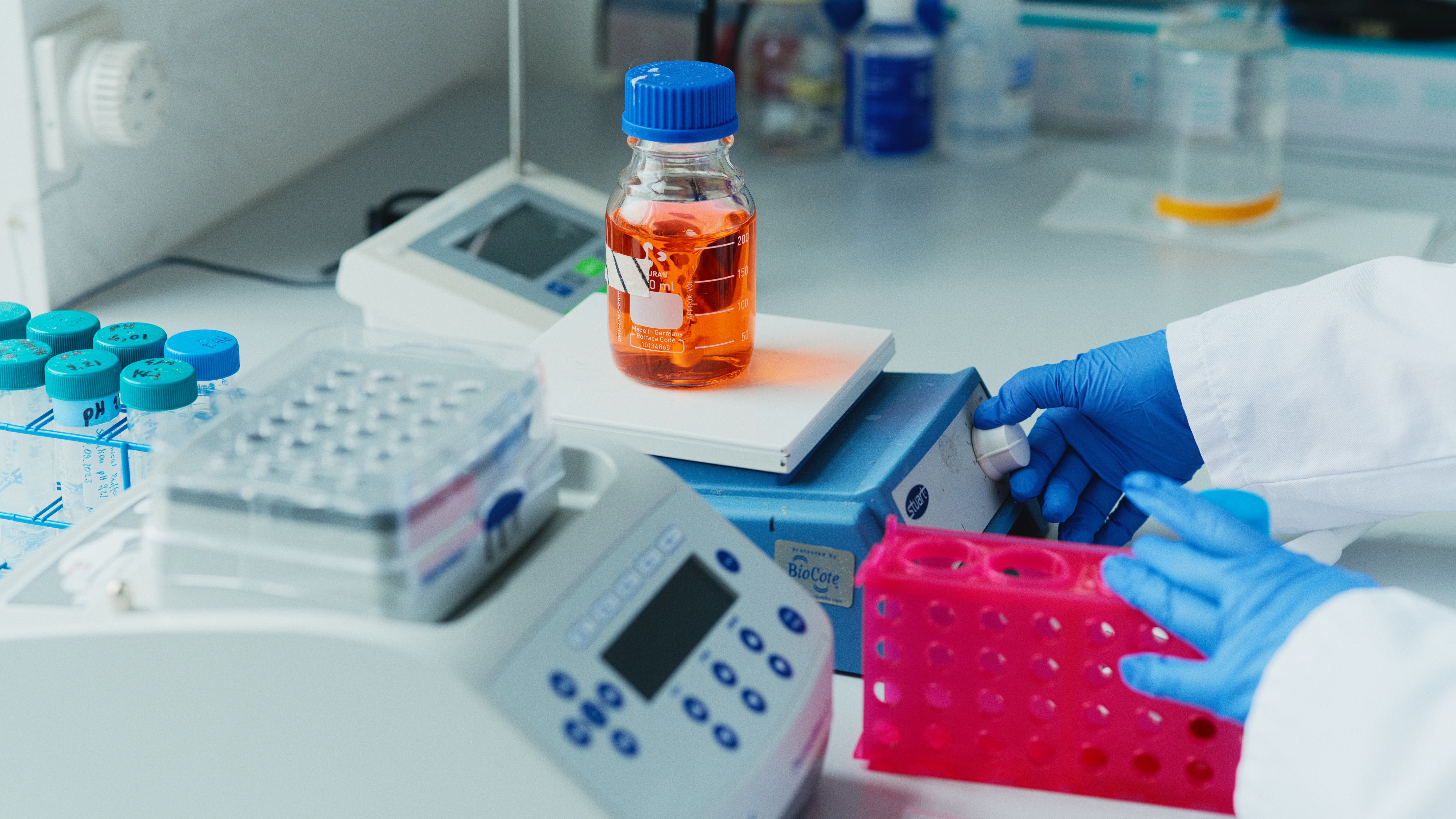 Ein Wissenschaftler in einem Labor trägt Schutzkleidung und Handschuhe. Vor ihm steht ein Waage mit einer Flasche orangefarbener Flüssigkeit. Neben der Waage befinden sich verschiedene Testbehälter und Reagenzgläser auf einem Tisch.
