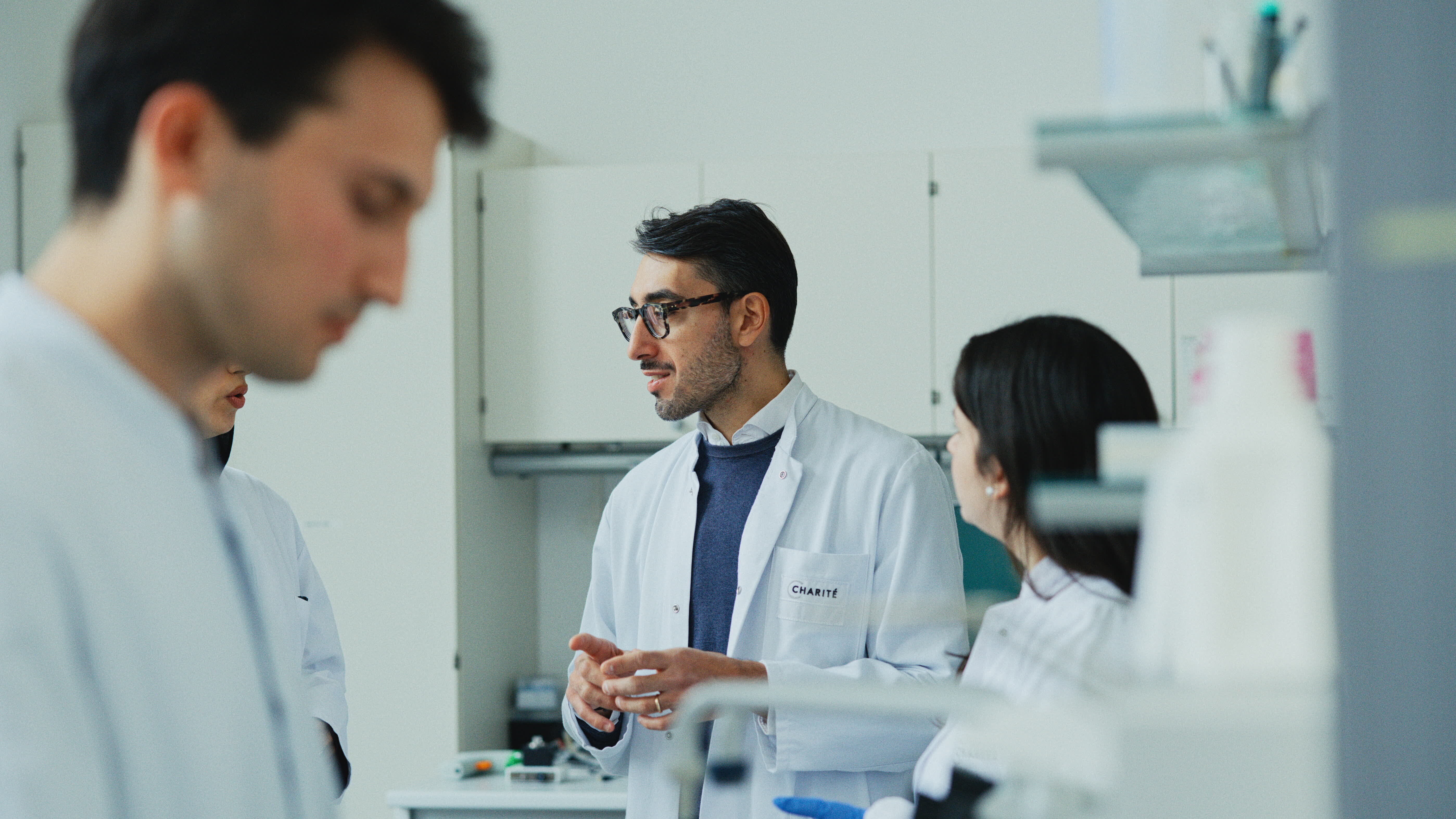 Vier Personen in Laborkleidung stehen in einem Labor. Ein Mann mit Brille erklärt etwas, während zwei andere Personen aufmerksam zuhören. Im Hintergrund ist ein Tisch mit Laborgeräten zu sehen. Alle wirken in einer professionellen und konzentrierten Arbeitsatmosphäre.