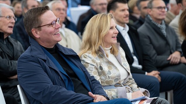 Eine Gruppe von Menschen sitzt in einem Publikum. Im Vordergrund sind ein Mann mit Brille und ein Lächeln sowie eine Frau in eleganter Kleidung zu sehen. Beide schauen interessiert und wirken in der Unterhaltung vertieft. Der Hintergrund zeigt weitere Personen, die ebenfalls aufmerksam sind.