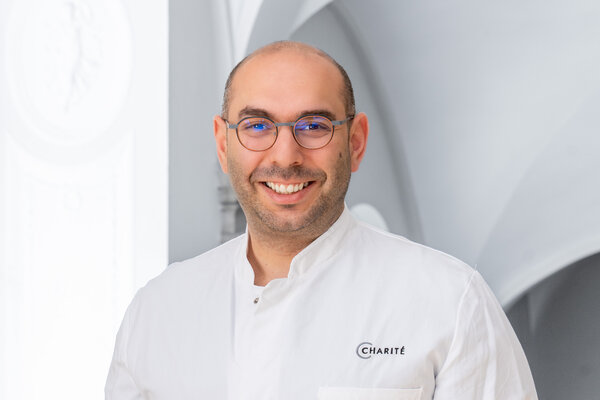 Ein lächelnder männlicher Arzt trägt einen weißen Kittel mit dem Logo der Charité. Er steht in einem hellen, architektonisch gestalteten Raum. Seine Brille hat einen dünnen Rahmen und er wirkt freundlich und einladend.