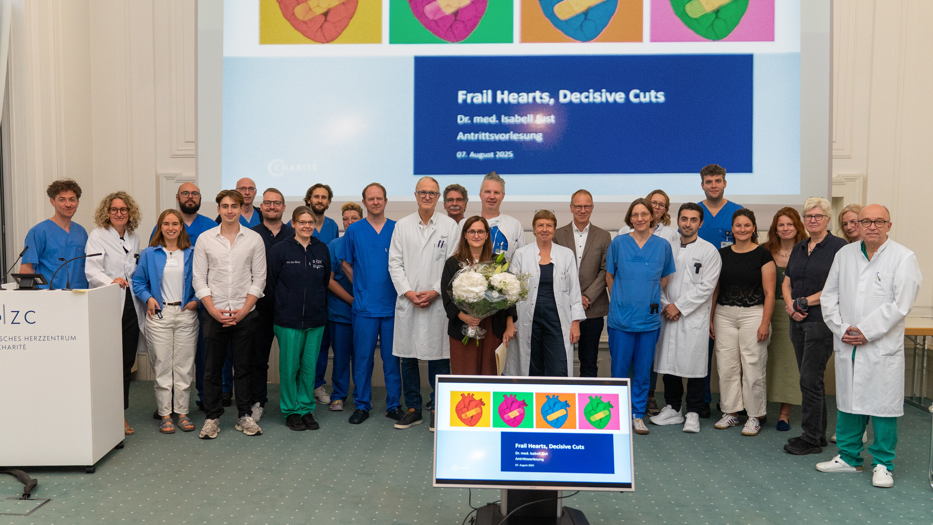 Eine Gruppe von Ärzten und Fachleuten steht zusammen in einem Raum, um eine Präsentation zum Thema "Frail Hearts, Decisive Cuts" zu halten. Im Hintergrund ist ein Bildschirm mit bunten Herzillustrationen und dem Titel der Vorlesung zu sehen. Eine Person hält einen Blumenstrauß.