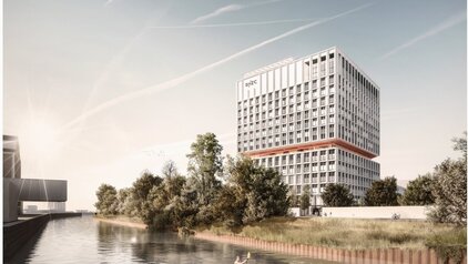 Eine moderne, mehrstöckige Gebäudestruktur steht nahe einem ruhigen Gewässer, umgeben von Bäumen und Grünflächen. Im Vordergrund ist ein gepflasterter Weg zu sehen, während im Wasser eine Person im Kanu paddelt. Der Himmel ist klar mit einigen Wolkenstreifen.