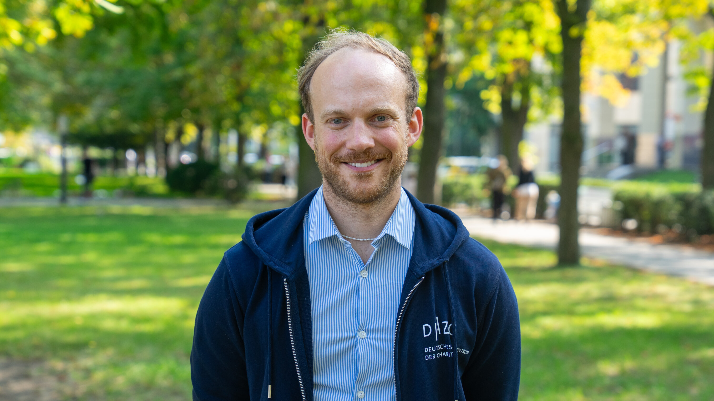 Ein junger Mann mit einem breiten Lächeln steht in einem Park. Er trägt ein blaues Sweatshirt und ein hellblaues, gestreiftes Hemd. Im Hintergrund sind grüne Bäume und verschwommen erkennbare Passanten zu sehen.