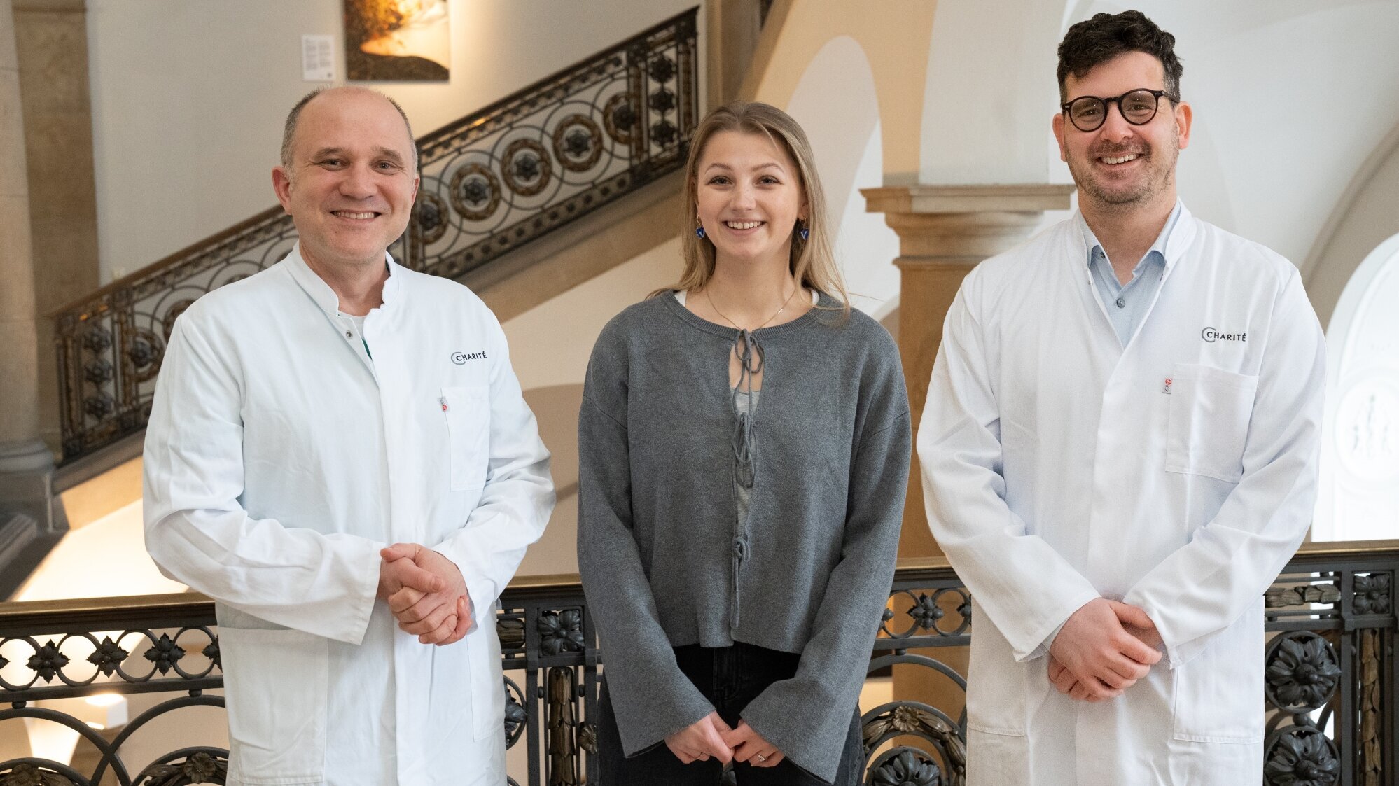 Drei Personen stehen in einem Gebäude: zwei Männer in weißen Laborkitteln und eine Frau in einem grauen Pullover. Sie lächeln und posieren vor einem eleganten Treppenabsatz mit Ornamenten. Die Szene vermittelt eine freundliche, professionelle Atmosphäre.