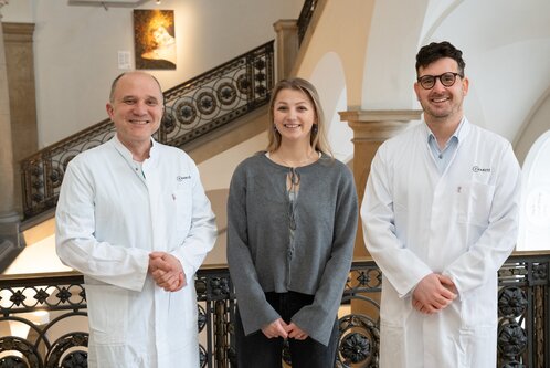 Drei Personen stehen in einem Gebäude: zwei Männer in weißen Laborkitteln und eine Frau in einem grauen Pullover. Sie lächeln und posieren vor einem eleganten Treppenabsatz mit Ornamenten. Die Szene vermittelt eine freundliche, professionelle Atmosphäre.