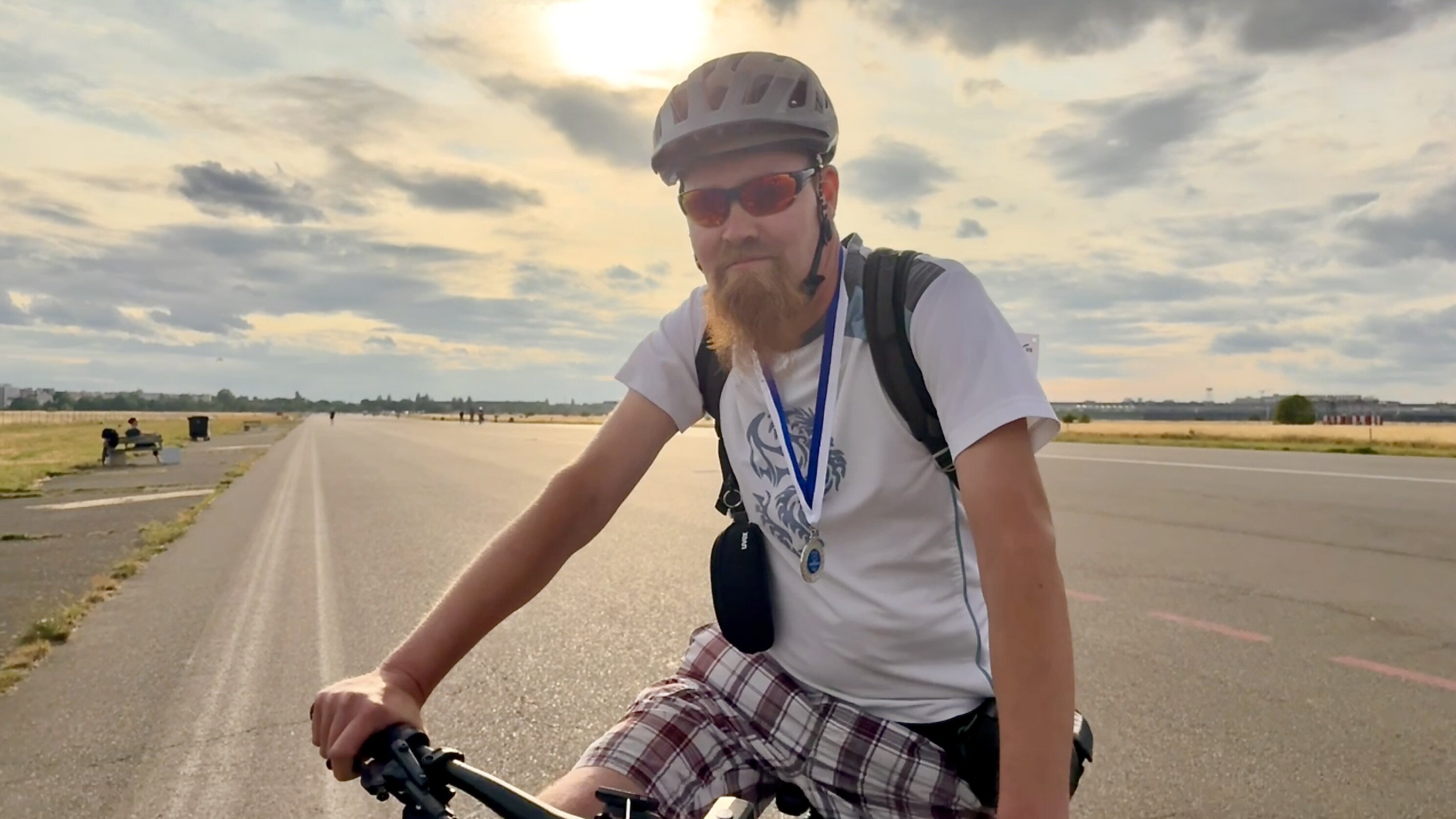Ein Mann mit Bart fährt auf einem Fahrrad. Er trägt einen Helm und eine Sonnenbrille. Um seinen Hals hängt eine Medaille. Im Hintergrund sind eine glatte Asphaltstraße und eine wolkige, helle Himmel zu sehen.