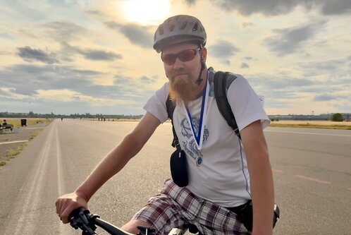 Ein Mann mit Bart fährt auf einem Fahrrad. Er trägt einen Helm und eine Sonnenbrille. Um seinen Hals hängt eine Medaille. Im Hintergrund sind eine glatte Asphaltstraße und eine wolkige, helle Himmel zu sehen.