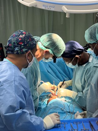 Operation im OP: Ein Team von Chirurgen in grüner und blauer Schutzkleidung führt einen Eingriff an einem Patienten durch, der auf einem Tisch liegt. Sie nutzen chirurgische Instrumente und konzentrieren sich auf den Operationsbereich.