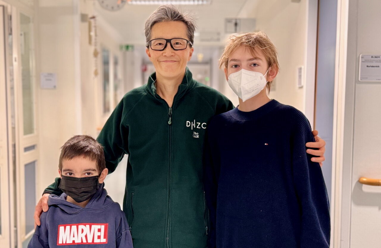 Ein Erwachsener steht in einem Krankenhausflur zwischen zwei Kindern. Alle drei tragen Mund-Nasen-Schutzmasken. Der Erwachsene trägt eine grüne Jacke, während die Kinder Sweatshirts mit unterschiedlichen Aufdrucken tragen. Der Flur wirkt hell und freundlich, mit medizinischen Ausstattungen im Hintergrund.