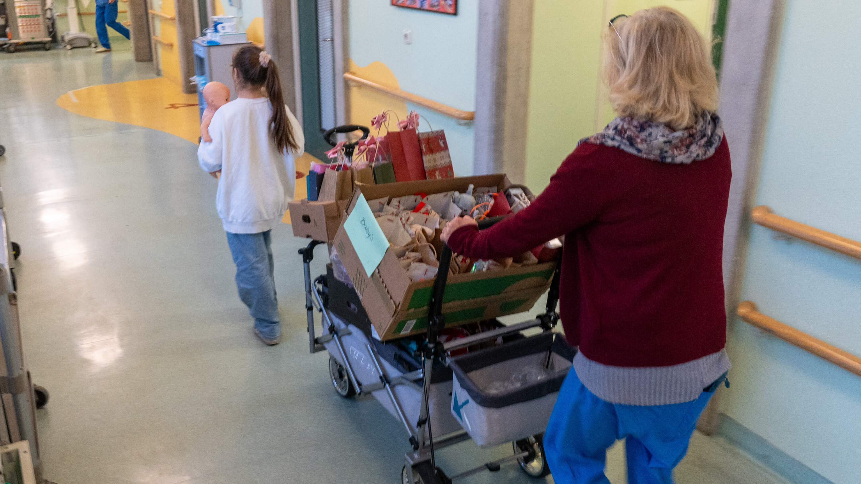 Eine Frau schiebt einen Wagen mit Geschenken durch einen Flur eines Krankenhauses. Ein Mädchen trägt eine Puppe und folgt ihr. Im Hintergrund sind medizinische Geräte und eine weitere Person in Praxisbekleidung sichtbar.