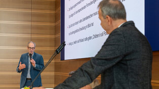 Ein Mann spricht an einem Rednerpult, während ein anderer Mann ihm zuhört. Im Hintergrund ist eine Präsentationsfolie mit Informationen über Kinder, Gesundheit und Politik zu sehen. Die Szene findet in einem modernen Konferenzraum statt.