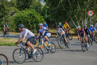 Eine Gruppe von Radfahrern fährt in einer Kurve auf einer Straße. Sie tragen Helme und sportliche Kleidung. Die Umgebung ist grün mit Bäumen im Hintergrund, und es gibt Verkehrsschilder in der Nähe. Einige Fahrer haben verschiedene Fahrradtypen.