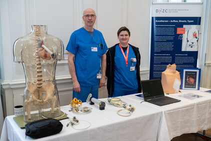 Zwei Personen in blauen Krankenhauskleidern stehen an einem Informationsstand. Neben ihnen befindet sich ein durchsichtiger Modellkörper, der einige innere Organe zeigt. Auf dem Tisch liegt ein Laptop und medizinische Geräte sowie Informationsmaterial zum Thema Kunstherzen.
