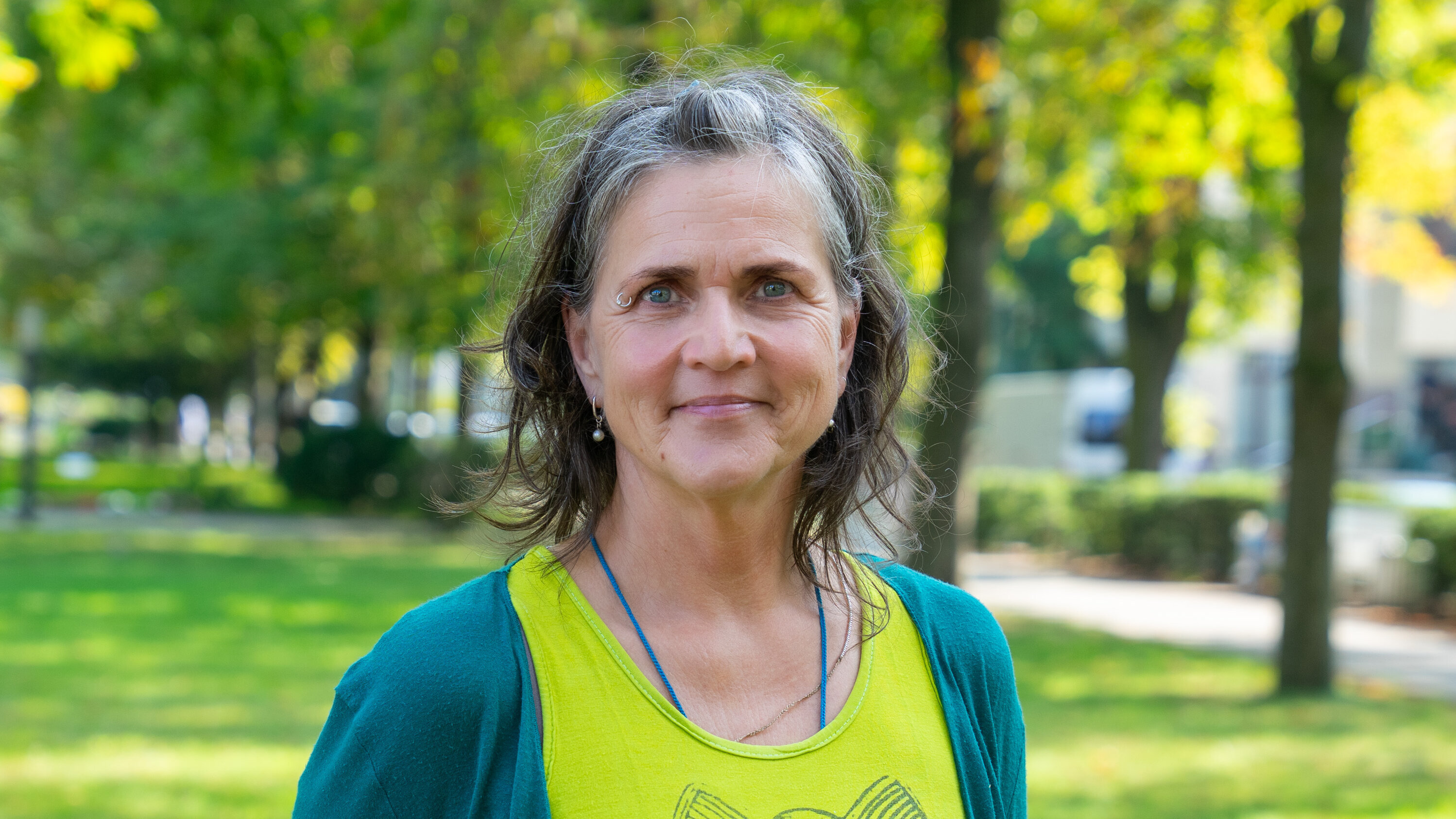 Eine lächelnde Frau mit langen, leicht gewellten Haaren steht im Freien. Sie trägt ein leuchtend grünes Oberteil mit einem Eulenmotiv und eine blaue Jacke. Im Hintergrund sind verschwommene Bäume und eine grüne Wiese zu erkennen.