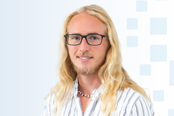 Ein junger Mann mit langen, blonden Haaren und einer Brille lächelt freundlich. Er trägt ein gestreiftes Hemd und eine Halskette. Der Hintergrund ist in sanften Blautönen mit geometrischen Formen.