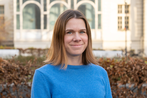Ein lächelnde Frau mit schulterlangem, brünettem Haar trägt einen blauen Pullover. Sie steht in einem Garten mit welken Blättern und einer Architektur im Hintergrund, die teilweise unscharf ist. Der Hintergrund vermittelt eine herbstliche Atmosphäre.