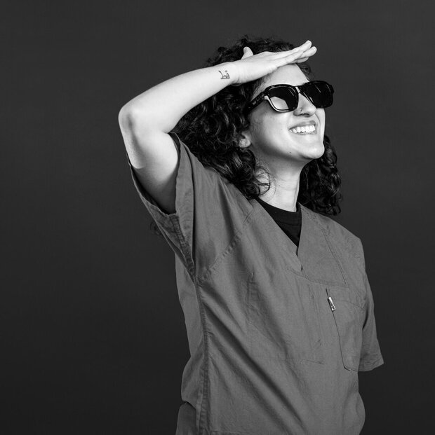 Eine fröhliche Person mit lockigem, schwarzem Haar trägt eine medizinische Kluft und Sonnenbrille. Sie steht mit einem Arm über der Augenbraue, als würde sie in die Ferne schauen. Der Hintergrund ist dunkel und sorgt für einen Kontrast zur Person im Vordergrund.