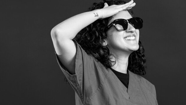 Eine fröhliche Person mit lockigem, schwarzem Haar trägt eine medizinische Kluft und Sonnenbrille. Sie steht mit einem Arm über der Augenbraue, als würde sie in die Ferne schauen. Der Hintergrund ist dunkel und sorgt für einen Kontrast zur Person im Vordergrund.