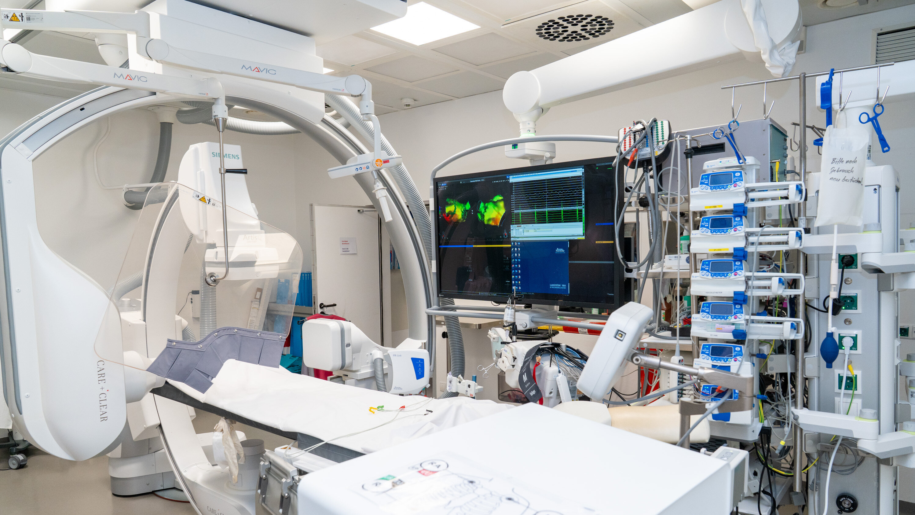 Das Bild zeigt einen modernen Operationssaal mit medizinischen Geräten. Im Vordergrund ist ein Operationsbett zu sehen, umgeben von einer Vielzahl von Monitoren und technischen Vorrichtungen, die für diagnostische und therapeutische Zwecke genutzt werden. Der Raum wirkt sauber und gut organisiert.