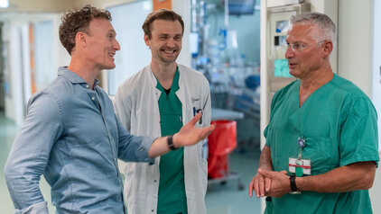 Drei Männer unterhalten sich in einem Krankenhausflur. Einer trägt ein hellblaues Hemd, der zweite einen weißen Arztkittel mit grüner Kleidung darunter und der dritte ist in grün gekleidet. Die Atmosphäre wirkt freundlich und informell.