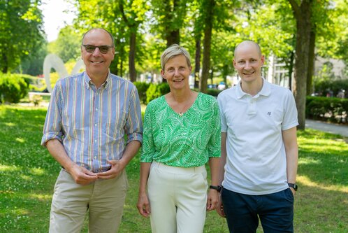 Drei Personen stehen im Freien auf einer grünen Wiese umgeben von Bäumen. Die Frau in der Mitte trägt ein grünes Oberteil, während die beiden Männer auf beiden Seiten lässige Hemden anhaben. Alle lächeln und wirken freundlich.