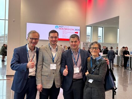 Vier Personen stehen in einem modernen Veranstaltungsraum und lächeln. Sie machen Daumen hoch. Im Hintergrund ist ein Banner mit der Aufschrift „HOTSPOT HERZMEDIZIN 2026“ zu sehen, sowie andere Besucher des Events.