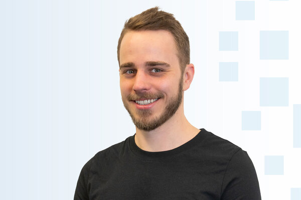 Ein junger Mann mit einem kurzen Bart und einem freundlichen Lächeln trägt ein schwarzes T-Shirt. Er steht vor einem hellen, strukturierten Hintergrund. Sein Gesichtsausdruck wirkt offen und einladend.