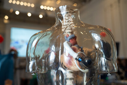 Das Bild zeigt ein transparentes Modell eines menschlichen Torso mit sichtbaren inneren Organen. In der Mitte befindet sich eine künstliche Herzpumpe. Der Torso ist in einer Umgebung mit unscharfen Hintergründen platziert, die für eine medizinische Demonstration oder Ausbildung genutzt wird.