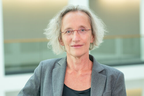Ein Porträt einer Frau mit grauen, lockigen Haaren, die einen grauen Blazer und ein schwarzes Oberteil trägt. Sie steht an einem Geländer in einem modernen Innenraum und schaut freundlich in die Kamera. Der Hintergrund ist hell und unscharf.