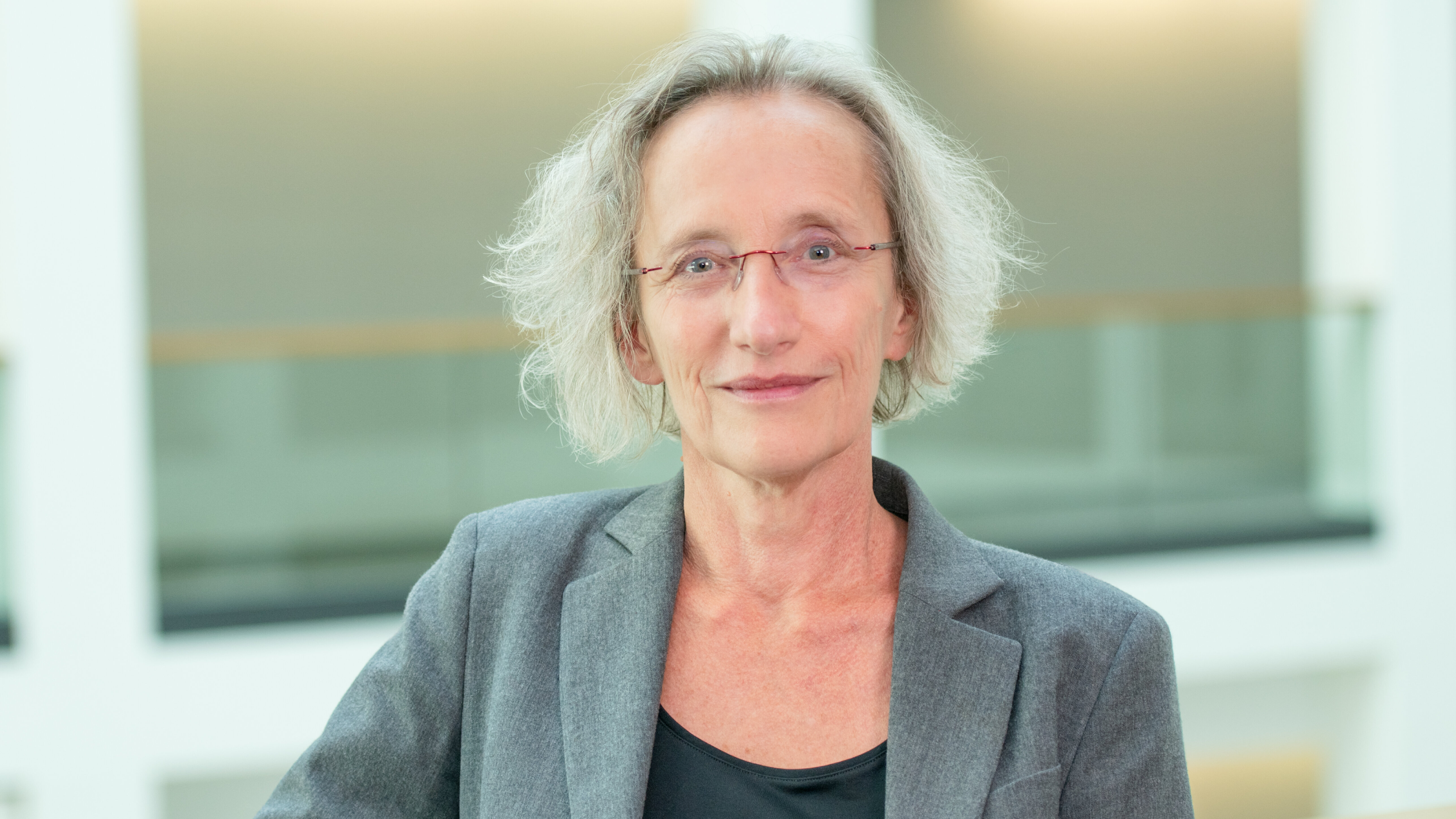 Ein Porträt einer Frau mit grauen, lockigen Haaren, die einen grauen Blazer und ein schwarzes Oberteil trägt. Sie steht an einem Geländer in einem modernen Innenraum und schaut freundlich in die Kamera. Der Hintergrund ist hell und unscharf.
