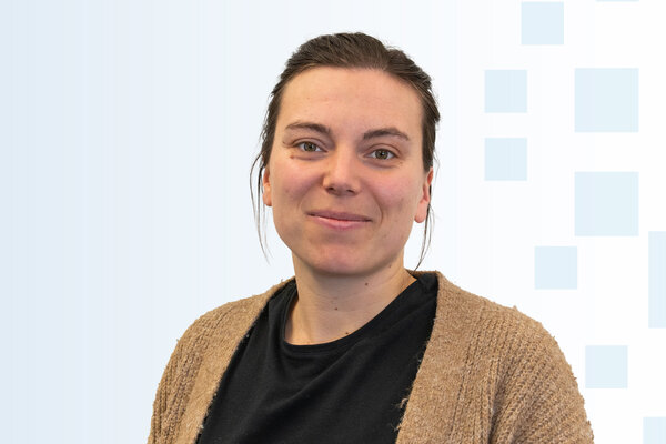 Porträt einer lächelnden Frau mit langen, dunklen Haaren. Sie trägt ein schwarzes T-Shirt und eine hellbraune Strickjacke. Der Hintergrund ist hell mit geometrischen Mustern in sanften Farben.