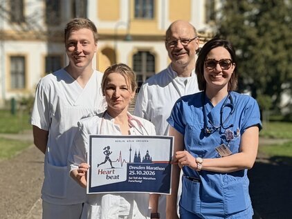 Vier Personen in medizinischer Kleidung stehen im Freien und halten ein Schild mit der Aufschrift „Heart Beat - Der Winter Marathon 25.10.2026“ in den Händen. Sie wirken freundlich und engagiert, die Umgebung scheint sonnig und einladend.