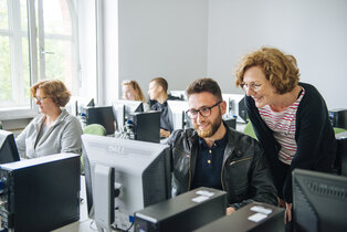 Eine Gruppe von fünf Personen in einem Computerraum. Zwei Personen im Vordergrund, ein Mann und eine Frau, betrachten den Bildschirm des Computers des Mannes. Die Frau lächelt, während der Mann freundlich reagiert. Im Hintergrund arbeiten drei weitere Personen an ihren Computern.