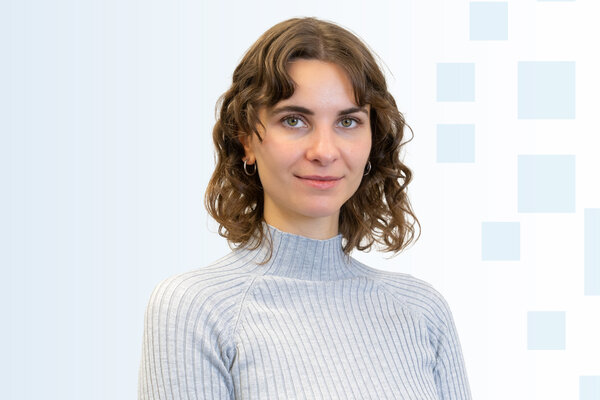 Porträt einer lächelnden Frau mit lockigem, braunem Haar, die ein hellgraues, geripptes Oberteil trägt. Der Hintergrund ist in sanften, hellen Farben mit geometrischen Mustern gestaltet. Die Frau schaut direkt in die Kamera und vermittelt eine freundliche Ausstrahlung.