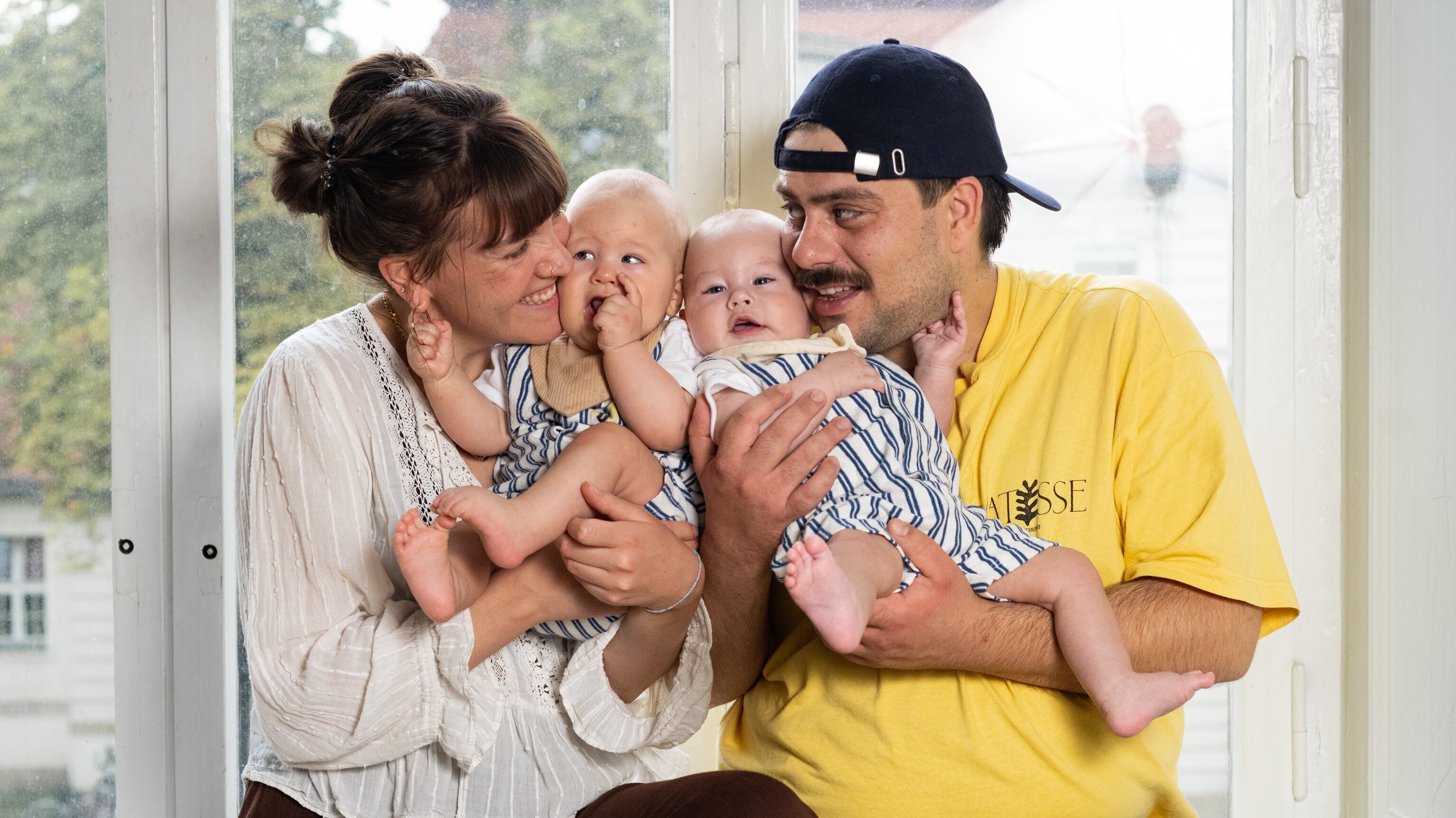 Eine Familie umarmt ihre Zwillinge. Die Mutter hält ein Baby auf dem Arm, während sie mit dem Vater lächelt, der das andere Baby hält. Die Szene vermittelt Freude und Zusammenhalt in einem hellen Raum mit Fensterblick.