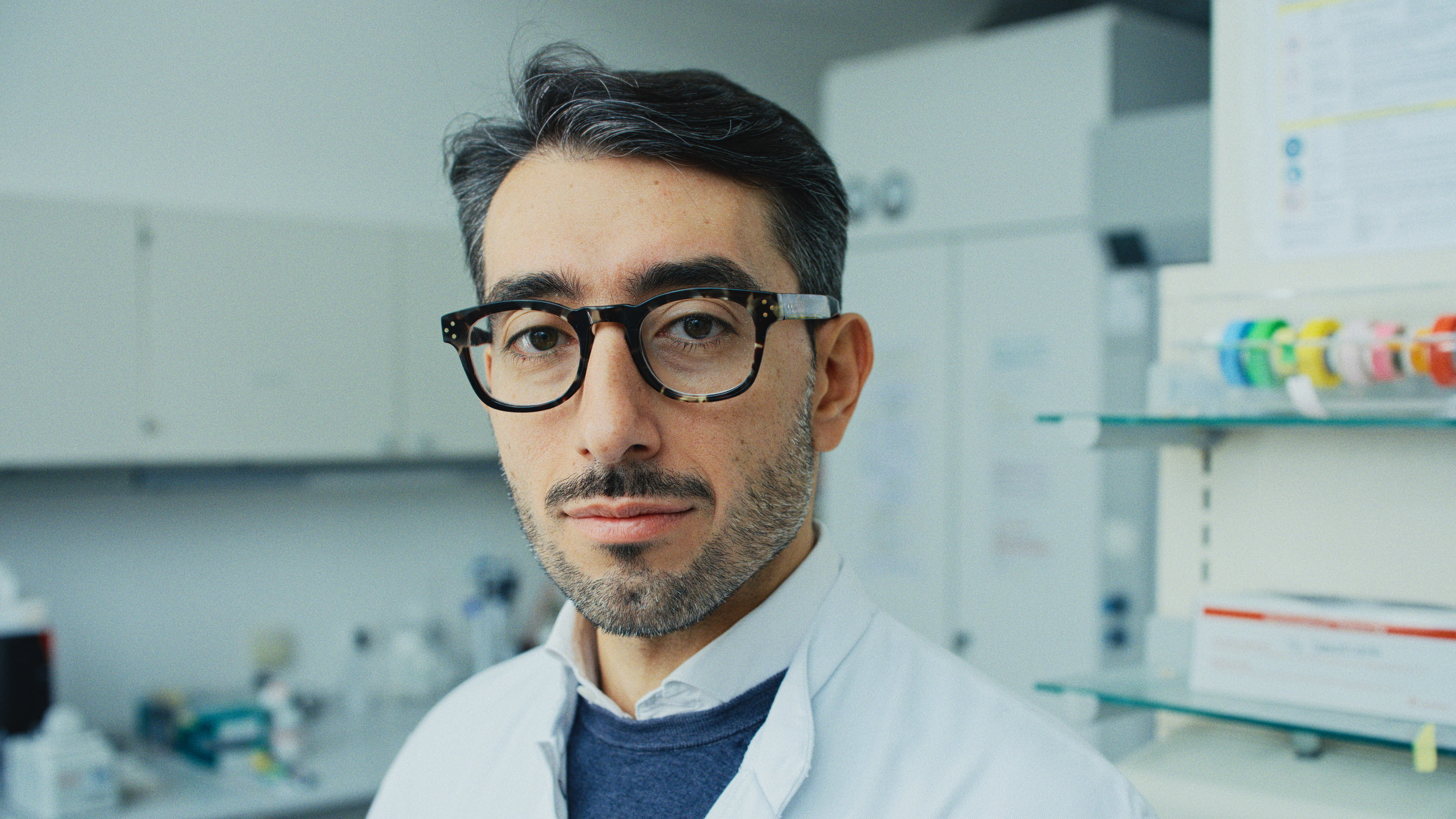 Ein Mann mit kurzen, dunklen Haaren trägt eine Brille und einen weißen Laborkittel. Er schaut in die Kamera und steht in einem modernen Labor mit Regalen im Hintergrund.