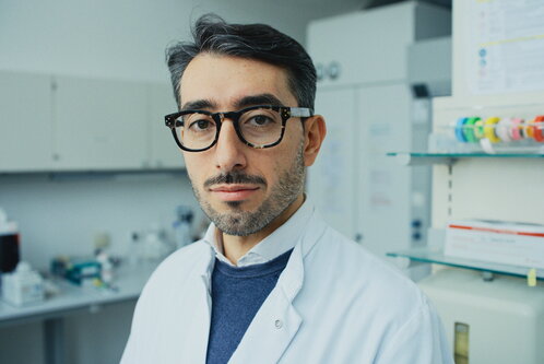 Ein Mann mit kurzen, dunklen Haaren trägt eine Brille und einen weißen Laborkittel. Er schaut in die Kamera und steht in einem modernen Labor mit Regalen im Hintergrund.