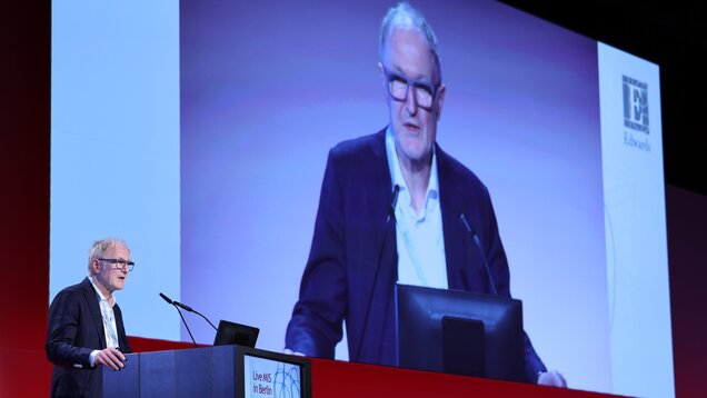 Ein Mann steht an einem Rednerpult und spricht in ein Mikrofon. Im Hintergrund ist sein Bild auf einer großen Leinwand zu sehen. Der Raum wirkt modern und professionell, wahrscheinlich während einer Konferenz oder Präsentation.