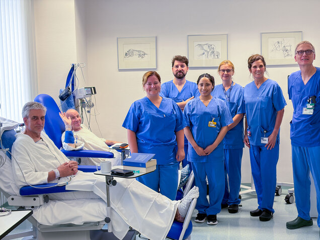 Eine Gruppe von sechs medizinischen Fachkräften in blauen Kitteln steht lächelnd zusammen. Vor ihnen sitzt ein Patient in einem weißen Nachthemd, der entspannt in einem Behandlungsstuhl sitzt. Die Umgebung wirkt freundlich und hell, was eine positive Atmosphäre vermittelt.