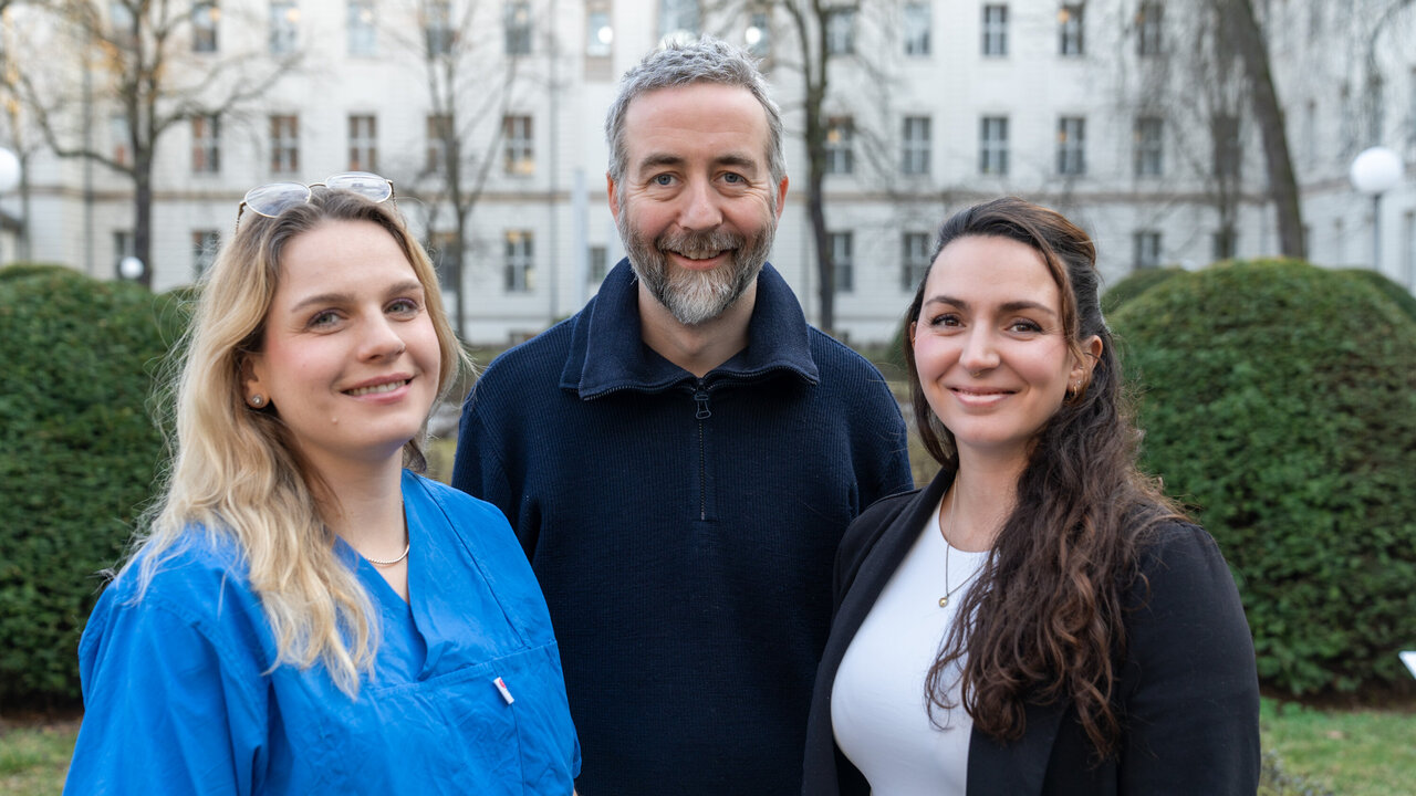 Drei Personen stehen im Freien vor einer hellen, historischen Gebäude-Fassade. Eine Frau trägt einen blauen Kittel, während die anderen beiden formelle Kleidung tragen. Alle drei lächeln und wirken freundlich. Das Bild vermittelt eine positive, kooperative Atmosphäre.