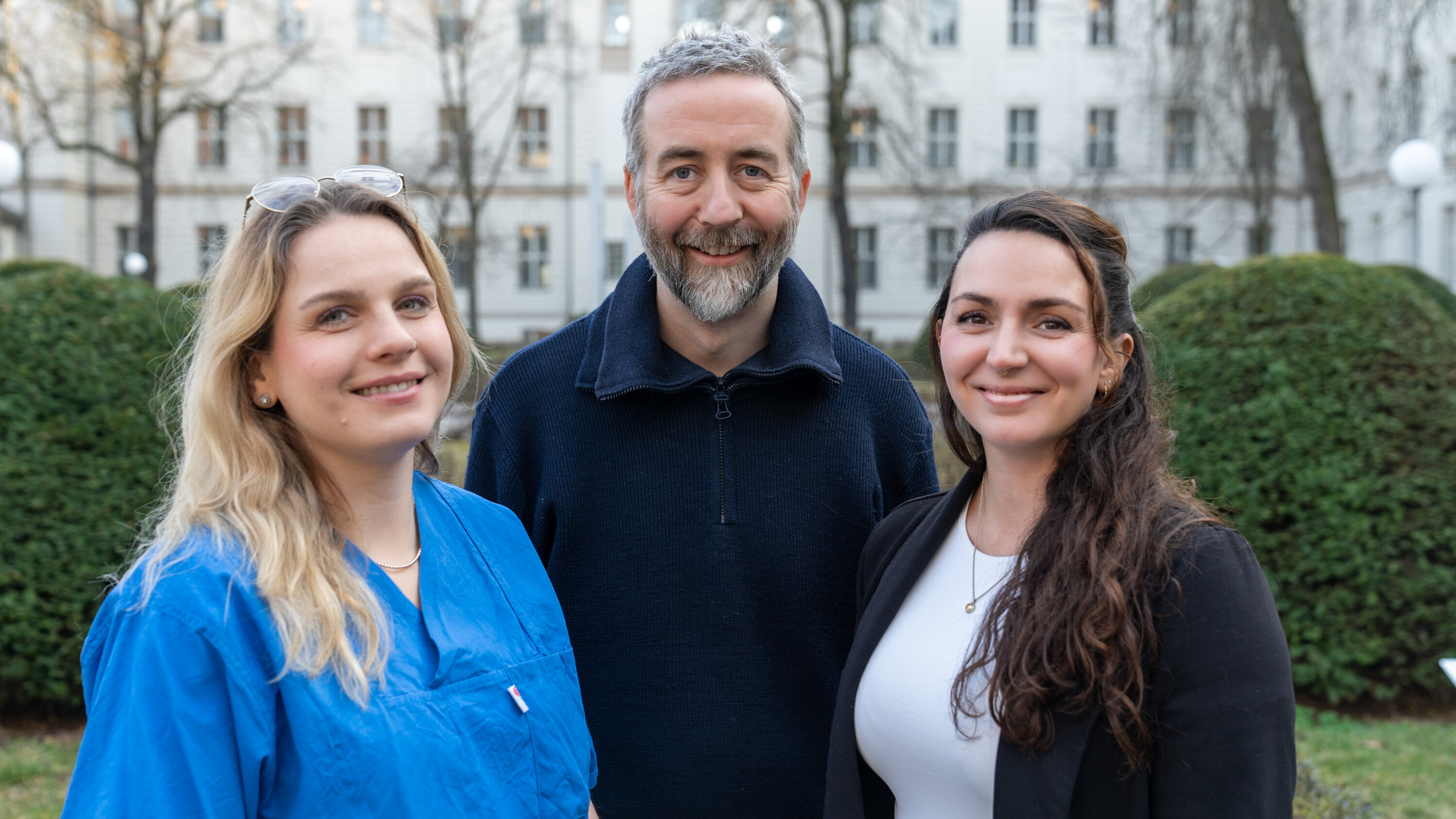 Drei Personen stehen im Freien vor einer hellen, historischen Gebäude-Fassade. Eine Frau trägt einen blauen Kittel, während die anderen beiden formelle Kleidung tragen. Alle drei lächeln und wirken freundlich. Das Bild vermittelt eine positive, kooperative Atmosphäre.