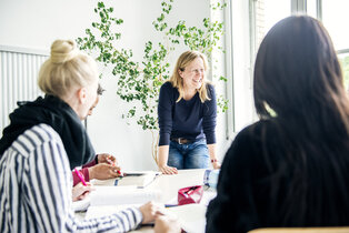 Eine lächelnde Frau steht an einem Tisch und spricht mit einer Gruppe von Menschen, die ihr aufmerksam zuhören. Einige Notizblätter und ein Buch sind auf dem Tisch sichtbar, während eine Pflanze im Hintergrund wächst.