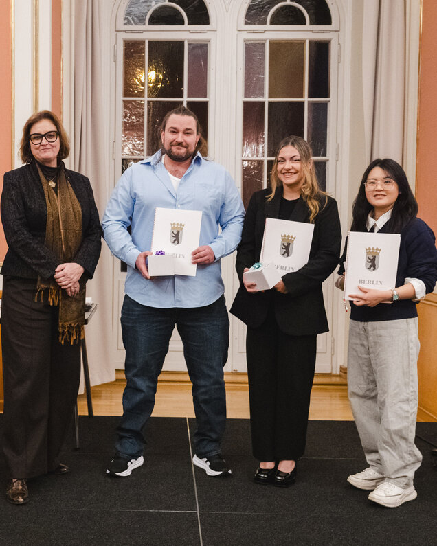 Vier Personen stehen auf einer Bühne in einem feierlichen Raum. Sie halten Urkunden mit dem Berliner Wappen in den Händen. Die Atmosphäre ist festlich, und die Personen lächeln. Im Hintergrund sind Fenster und eine dekorative Wand zu sehen.