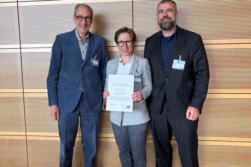 Drei Personen stehen nebeneinander vor einer Holzpaneelwand. Die mittlere Person hält ein Zertifikat in der Hand und trägt einen grauen Blazer. Die beiden anderen Personen stehen links und rechts davon, lächeln und sind formell gekleidet.