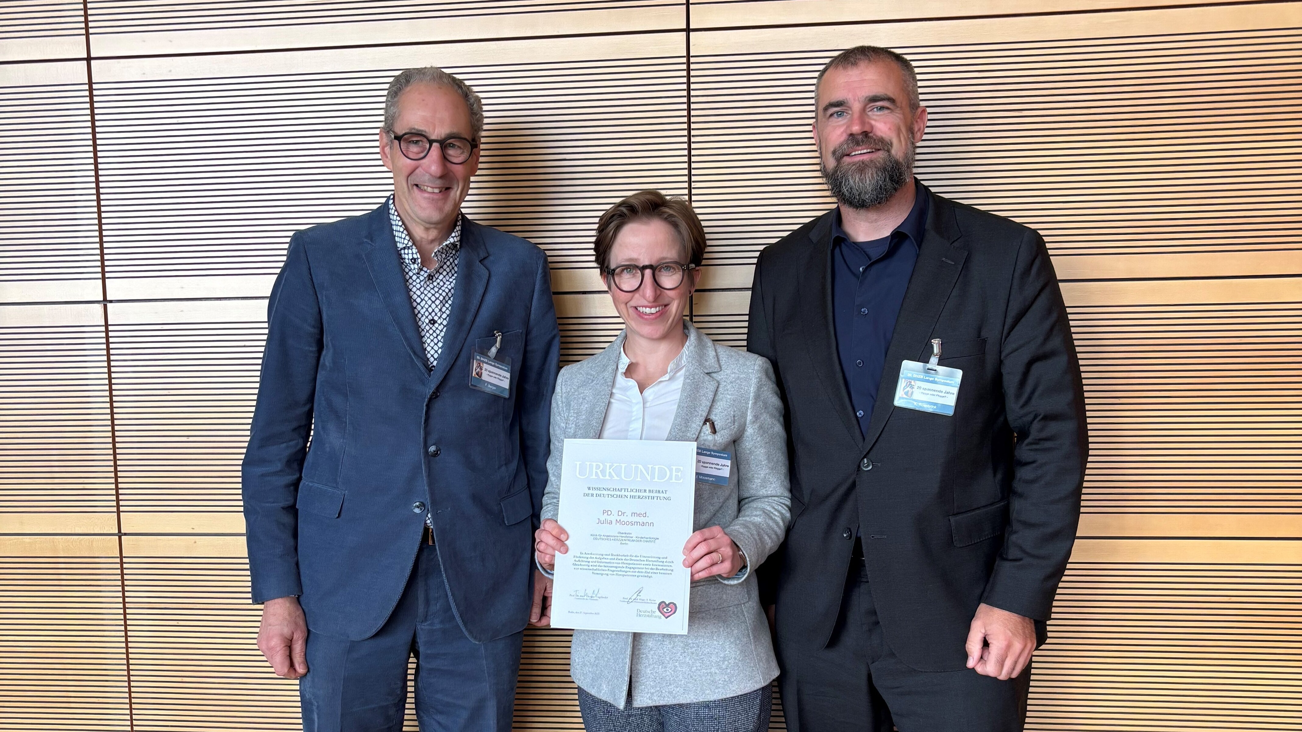 Drei Personen stehen nebeneinander vor einer Holzpaneelwand. Die mittlere Person hält ein Zertifikat in der Hand und trägt einen grauen Blazer. Die beiden anderen Personen stehen links und rechts davon, lächeln und sind formell gekleidet.