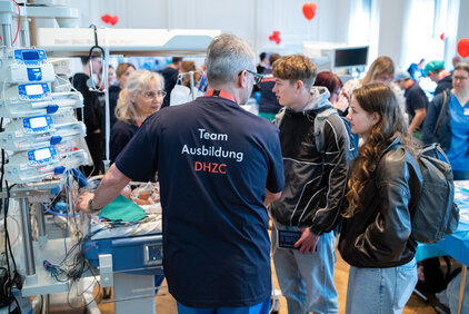 Eine Person mit einem schwarzen T-Shirt, auf dem "Team Ausbildung DHZC" steht, erklärt einer Gruppe junger Menschen etwas an einem medizinischen Gerät. Der Raum ist hell erleuchtet und es sind weitere Personen im Hintergrund sichtbar.