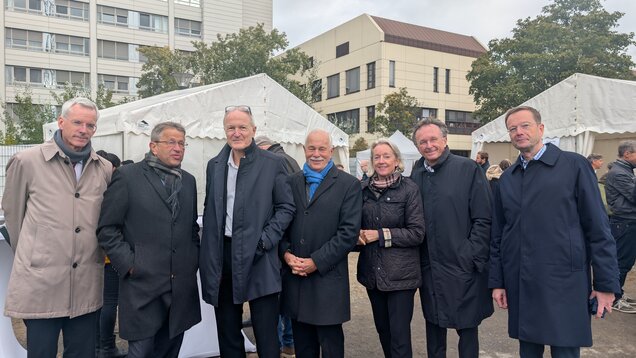 Eine Gruppe von sieben Personen steht im Freien, alle tragen dunkle Mäntel. Sie lächeln und scheinen an einer Veranstaltung teilzunehmen. Im Hintergrund sind Zelte und ein modernes Gebäude zu sehen.