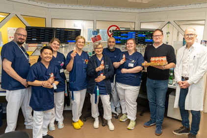 Eine Gruppe von neun Personen in blauen Uniformen steht in einem Raum voller Bildschirme. Mehrere Mitglieder halten Schokoladenfiguren in der Hand, während ein Mann in Zivil einen Tablett mit weiteren Schokoladenfiguren präsentiert. Alle lächeln und scheinen eine positive Atmosphäre zu haben.