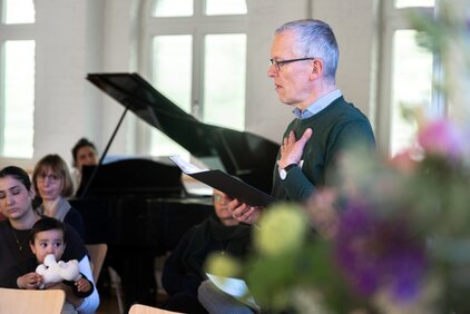 Ein älterer Mann mit Brille spricht gefühlvoll zu einer Gruppe von Zuhörern, darunter eine Frau mit einem Kind. Der Mann hält ein Blatt Papier in der Hand und ist an einer musikalischen Umgebung, möglicherweise in einem Veranstaltungsraum, zu sehen.